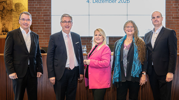 Fünf Personen stehen nebeneinander vor einer Präsentationswand, auf der „Verbandsversammlung“ und das Datum „4. Dezember 2025“ steht. Die Personen sind formell gekleidet, mit einem Fokus auf einer Dame in einem pinken Blazer.