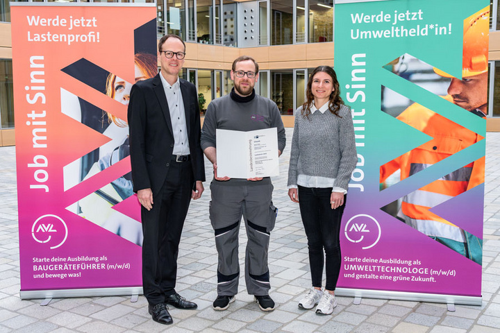 Drei Personen stehen in einem Innenhof. Eine Person hält ein Dokument in der Hand. Im Hintergrund sind zwei Banner zu sehen, die für Ausbildungsangebote in den Bereichen Baugeräteführung und Umwelttechnologie werben. Es wird zur Teilnahme an diesen Ausbildungsberufen aufgerufen.