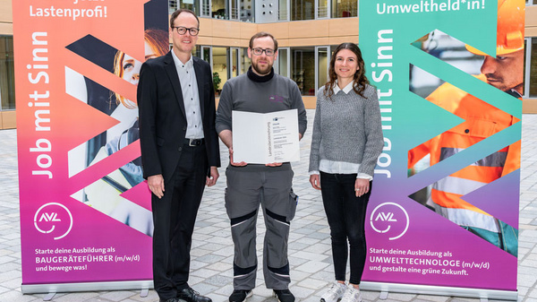 Drei Personen stehen in einem Innenhof. Eine Person hält ein Dokument in der Hand. Im Hintergrund sind zwei Banner zu sehen, die für Ausbildungsangebote in den Bereichen Baugeräteführung und Umwelttechnologie werben. Es wird zur Teilnahme an diesen Ausbildungsberufen aufgerufen.