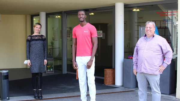 Three individuals stand outside a building. A woman in a black and gray dress is on the left, a man in a red shirt and white pants is in the center, and a man in a purple shirt is on the right. They appear to be posing for a photo.