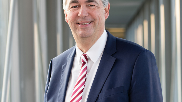 A smiling man is dressed in a dark suit and a white shirt with a striped tie. He stands confidently in a modern corridor, with glass walls and a well-lit background, conveying a professional and approachable demeanor.