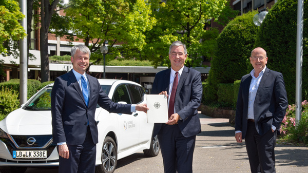 Three men stand outdoors near a white electric car. One man in a suit hands over a document to another, who appears to be presenting it. The background features greenery and a building, indicating a formal event likely related to business or collaboration.