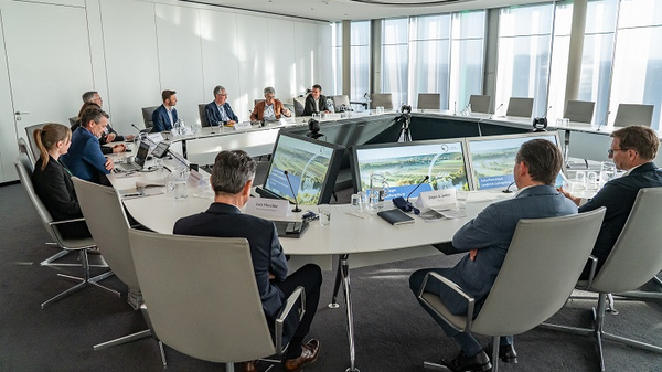 Eine Gruppe von Fachleuten sitzt an einem großen, runden Tisch in einem modernen Konferenzraum. Mehrere Personen sind in Vorstellungsgespräche verwickelt, während große Bildschirme am Tisch Präsentationen zeigen. Die Atmosphäre wirkt geschäftlich und konzentriert.