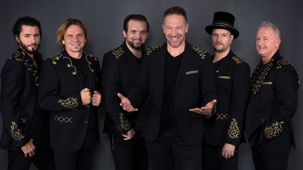 Un gruppo di sei musicisti è in posa, vestiti con giacche nere decorate da ricami dorati. Il membro centrale sorride e apre le braccia, mentre gli altri si presentano con espressioni amichevoli. Lo sfondo è scuro, creando un contrasto con i loro abiti.