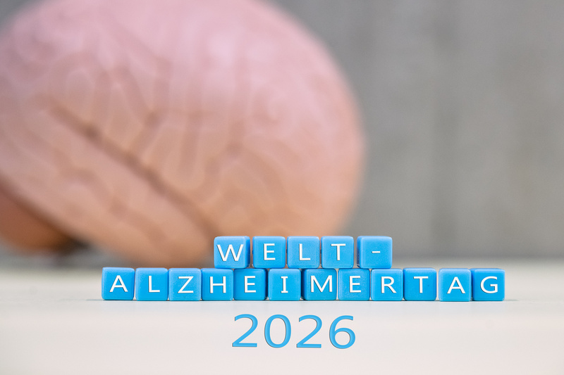 Blaue Würfel mit den aufgedruckten Worten "WELT-ALZHEIMERTAG" und "2026" im Vordergrund, im Hintergrund eine stilisierte Darstellung eines menschlichen Gehirns. (Text KI generiert)