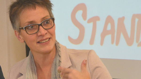 Eine Frau mit kurzen, grauen Haaren und Brille sitzt an einem Tisch. Sie trägt einen blassen Blazer und hat einen Schal um den Hals. Sie gestikuliert mit der rechten Hand und spricht anscheinend leidenschaftlich. Im Hintergrund steht das Wort "STANDPUNKT".