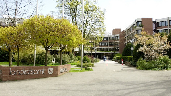 A courtyard scene featuring the entrance to a government building, marked by a sign that reads "landratsamt." Surrounding the entrance are green trees and shrubs, with people walking along the path. The setting is bright and welcoming, indicating a public space.