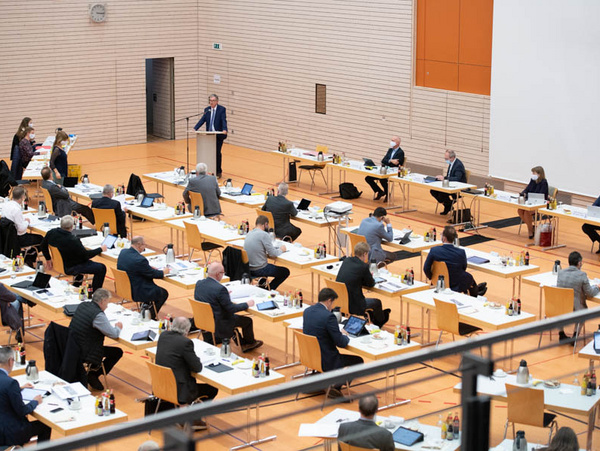 23.10.2020 - Kreistagssitzung in Oberstenfeld-Gronau Eine Sitzung in einem Konferenzraum mit mehreren Tischen, an denen zahlreiche Teilnehmende sitzen. Ein Redner steht vorne am Pult. Die Anwesenden sind in Diskussion vertieft und nutzen elektronische Geräte. Der Raum hat eine helle, moderne Atmosphäre.
