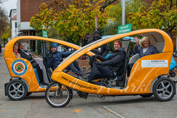 Zwei bunte, elektrisch betriebene Fahrradrikschas stehen nebeneinander auf einer Straße. In jeder Rikschafahren mehrere Personen, die fröhlich zur Kamera winken. Im Hintergrund sind herbstliche Bäume zu sehen. Ein Schild weist auf Klimaschutz hin.