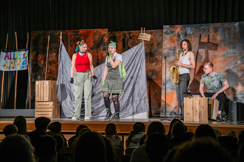 Zwei Schauspielerinnen in auffälligen Kostümen stehen auf einer Bühne und sprechen miteinander. Im Hintergrund sind Kulissen mit Farben und Texten. Ein Musiker spielt Saxophon, während eine weitere Person auf einer Kiste sitzt. Das Publikum ist im Vordergrund zu sehen. (Text KI generiert)