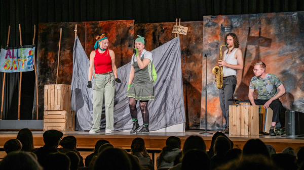 Zwei Schauspielerinnen in auffälligen Kostümen stehen auf einer Bühne und sprechen miteinander. Im Hintergrund sind Kulissen mit Farben und Texten. Ein Musiker spielt Saxophon, während eine weitere Person auf einer Kiste sitzt. Das Publikum ist im Vordergrund zu sehen. (Text KI generiert)