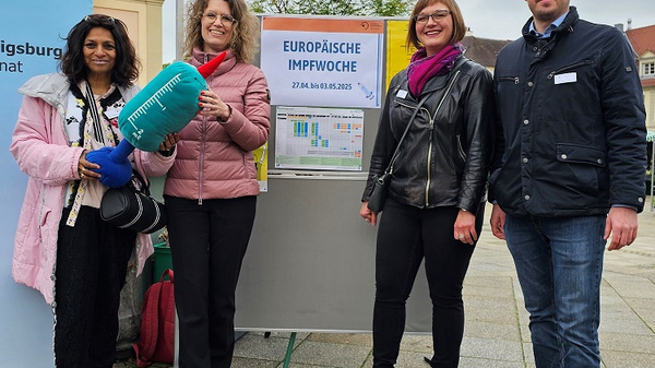 Vier Personen stehen an einem Stand, der für die Europäische Impfwoche wirbt. Eine Frau hält eine große Spritze, während die anderen in lässiger Kleidung lächeln. Im Hintergrund ist ein Plakat mit dem Datum der Impfwoche zu sehen. Es herrscht eine freundliche, informative Atmosphäre.