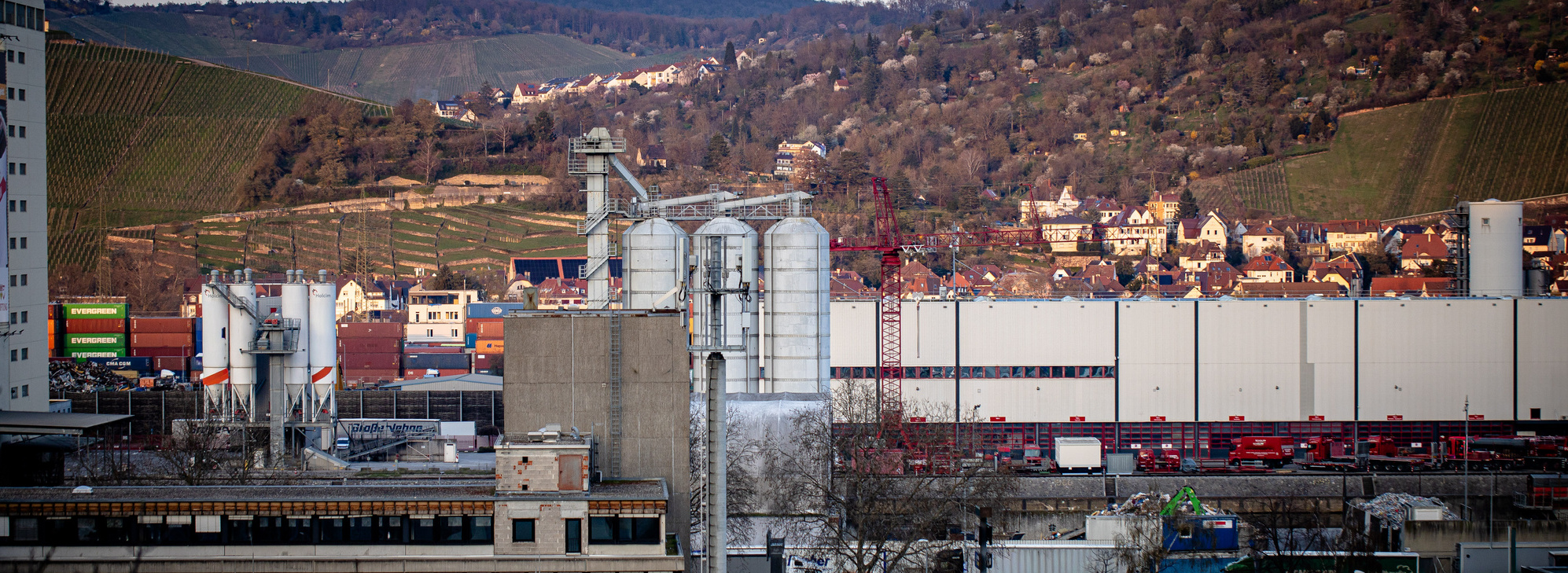 Header Bild: Industrieansicht mit Silos im Vordergrund, im Hintergrund eine bergige Landschaft und ein Wohngebiet. Die Szene zeigt die Verbindung von urbaner und natürlicher Umgebung, wobei die industrielle Architektur dominiert.