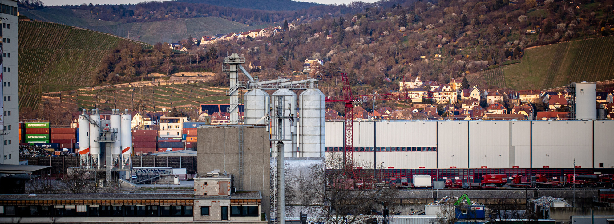 Header Bild: Industrieansicht mit Silos im Vordergrund, im Hintergrund eine bergige Landschaft und ein Wohngebiet. Die Szene zeigt die Verbindung von urbaner und natürlicher Umgebung, wobei die industrielle Architektur dominiert.