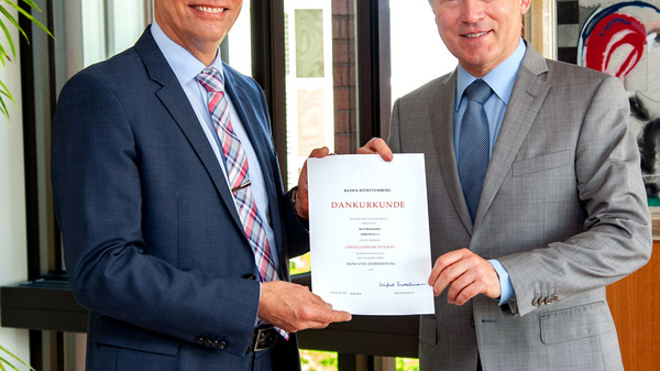 Two men stand side by side, one holding a certificate that reads "DANKURUNDE." They share smiles in a well-lit office setting, with greenery visible in the background. The scene conveys a sense of accomplishment and recognition.