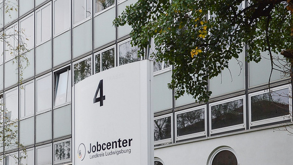 A modern building features large glass windows. In front, a prominent sign displays the number "4" and the word "Jobcenter," indicating the location of a job center in Lüneburg. Surrounding trees add greenery to the urban setting.