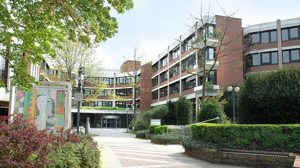 A modern building complex with three stories features a welcoming entrance surrounded by greenery and landscaped areas. The path leading up to the entrance is lined with trees and bushes, creating an inviting atmosphere. A colorful mural is visible on the left side.