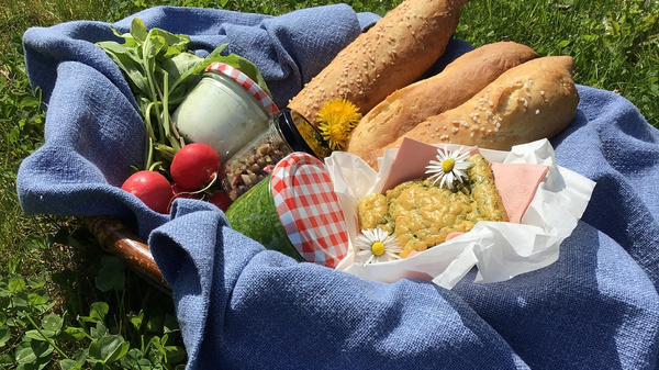 A picnic basket lined with a blue cloth holds freshly baked bread, vibrant radishes, a jar of yogurt, and a green cucumber. A slice of cake, topped with daisies, is wrapped in paper. The background features green grass, evoking a pleasant outdoor setting.