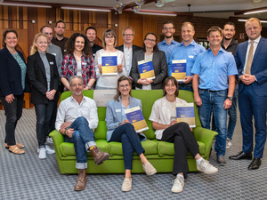 A diverse group of individuals, including men and women of various ages, stand and sit on a green couch, smiling and holding certificates. The setting is a modern indoor space with wooden paneling. The atmosphere is celebratory, indicating an achievement or recognition event.