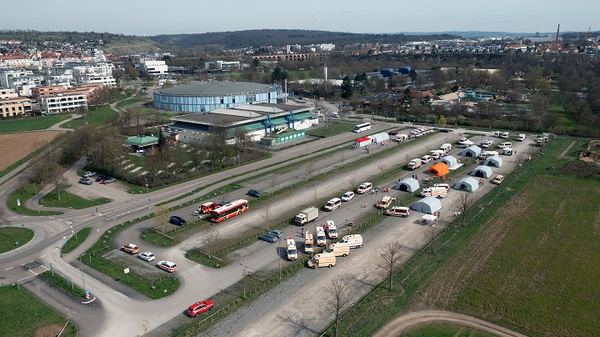 Luftaufnahme eines Parkplatzes mit mehreren Fahrzeugen und Zelten. Im Hintergrund befindet sich ein modernes Gebäude, umgeben von grünen Flächen und weiteren Gebäuden. Die Szene vermittelt einen Eindruck von Aktivität und Organisation in der Umgebung.