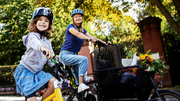 Due donne sorridenti in bicicletta, una con un casco e una maglietta grigia, l'altra su una bici con un rimorchio. Sono circondate da alberi e un ambiente all'aperto, mostrando gioia e divertimento in un percorso ciclabile.