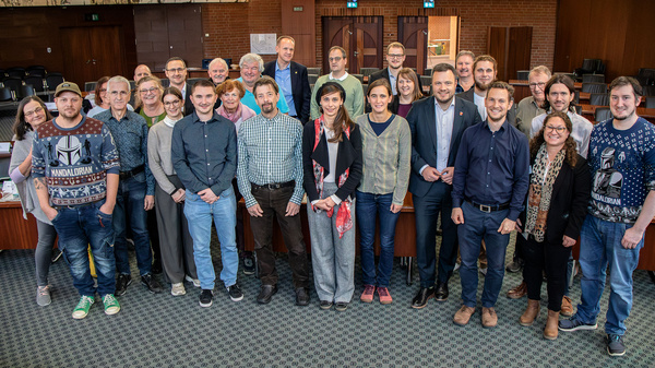 Eine Gruppe von etwa 30 Personen steht in einem Konferenzraum. Sie schauen in die Kamera und lächeln. Hinter ihnen sind Stühle und große Fenster sichtbar. Die Anwesenden tragen verschiedene Kleidung, darunter formelle und legere Outfits.