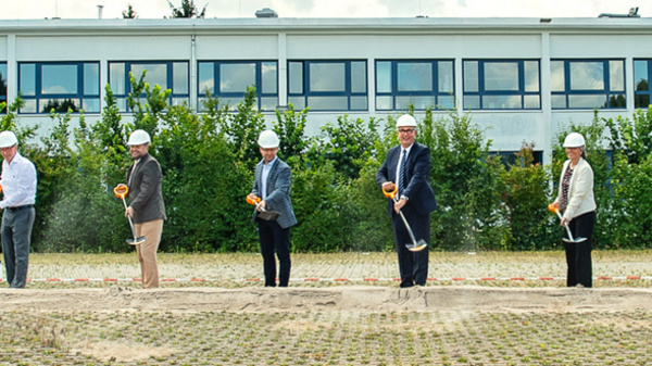 A group of professionals in hard hats stands in a line, each holding a shovel, in front of a building and trees. They appear to be participating in a ground-breaking ceremony, symbolizing the start of a construction or development project.