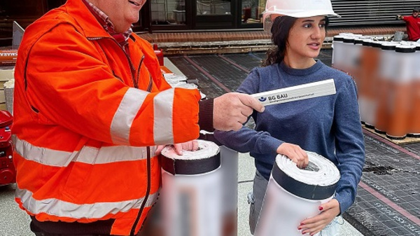 Zwei Personen auf einer Baustelle: Ein Mann mit orangefarbener Warnjacke und Helm zeigt mit einem Maßstab auf ein Rohr, während eine junge Frau in blauer Kleidung daneben steht und ein weiteres Rohr hält. Im Hintergrund sieht man einen Gebäudekomplex.