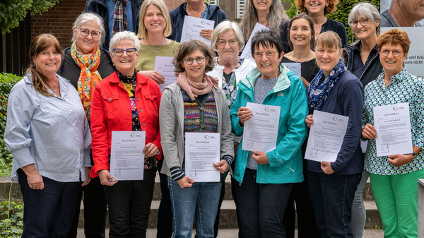 Un groupe de 15 personnes se tient à l'extérieur, tenant des documents. Elles affichent des sourires et semblent engagées dans une activité liée à la sensibilisation à la démence. En arrière-plan, une affiche affiche des informations sur les soins pour les personnes atteintes de démence.