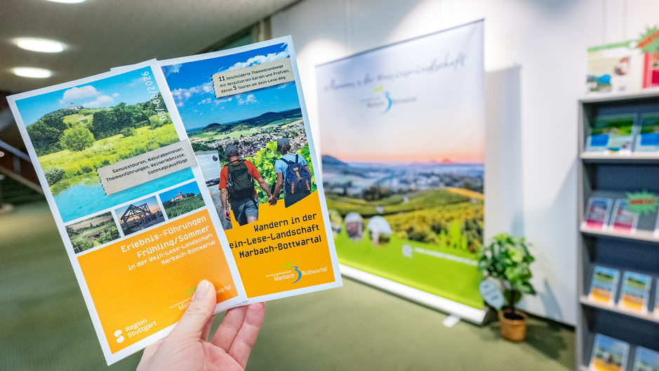 Una mano regge un opuscolo con informazioni sul Bottwartal. Sullo sfondo c'è uno stand con una foto del Bottwartal.