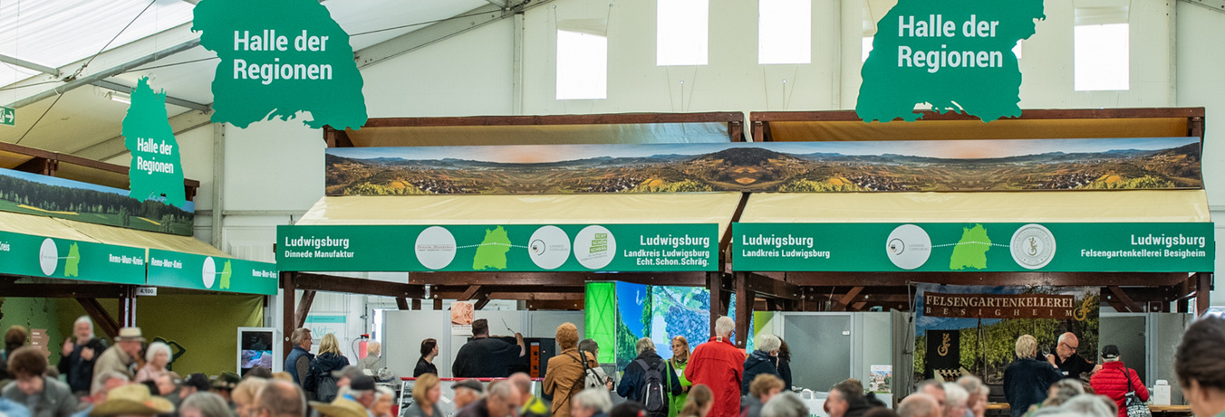In einer großen Halle mit mehreren Ständen stehen zahlreiche Menschen zusammen. Über den Ständen hängen Schilder mit der Aufschrift „Halle der Regionen“. Die Atmosphäre ist lebhaft und belebend, wobei verschiedene regionale Produkte und Informationen angeboten werden. (Text KI generiert)