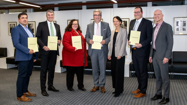 Eine Gruppe von sechs Personen steht in einem Raum und hält jeweils ein gelbes Dokument in den Händen. Sie zeigen sich freundlich und stehen in einer Reihe. Der Raum hat eine moderne Einrichtung mit Bildern an der Wand.