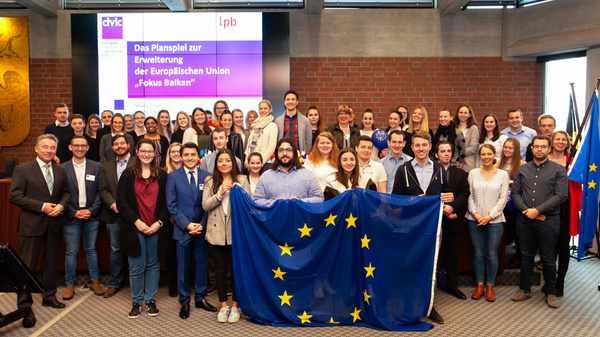 Un gruppo di circa cinquanta persone partecipa a un evento, sorreggendo una grande bandiera dell'Unione Europea. Sullo sfondo, un proiettore mostra un titolo riguardante l'allargamento dell'Unione Europea, con il tema "Fokus Balkan". L'atmosfera è coinvolgente e collaborativa.