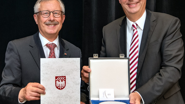 Deux hommes sont en train de poser pour une photo. L'un tient un document avec un blason, tandis que l'autre présente une médaille dans une boîte. Les deux sourient, dans un cadre formel.