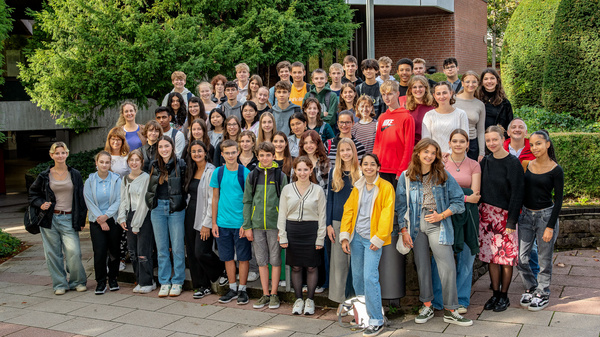 Eine große Gruppe von Schülern steht im Freien zusammen. Sie sind unterschiedlichen Alters und Geschlechts, tragen verschiedene Kleidung und lächeln freundlich in die Kamera. Im Hintergrund sind Bäume und ein Gebäude sichtbar.