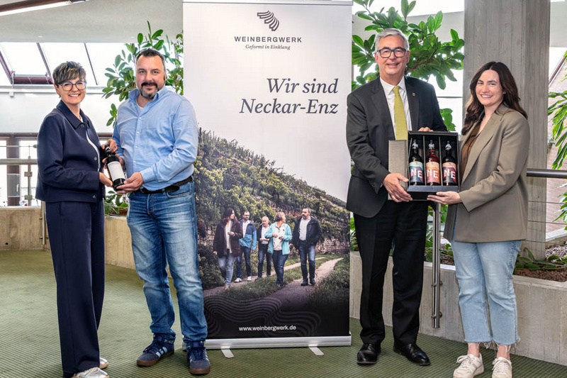 Vier Personen stehen zusammen vor einem Banner mit der Aufschrift „Wir sind Neckar-Enz“. Zwei Personen halten eine Flasche, während die anderen zwei eine Holzkiste mit mehreren Flaschen präsentieren. Die Szene spielt in einem modernen Innenraum mit Pflanzen im Hintergrund.