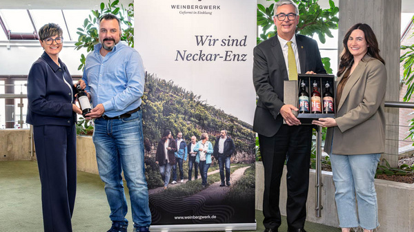 Vier Personen stehen zusammen vor einem Banner mit der Aufschrift „Wir sind Neckar-Enz“. Zwei Personen halten eine Flasche, während die anderen zwei eine Holzkiste mit mehreren Flaschen präsentieren. Die Szene spielt in einem modernen Innenraum mit Pflanzen im Hintergrund.