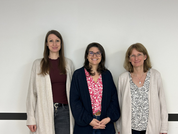 Drei Frauen stehen nebeneinander vor einer hellen Wand. Die Frau in der Mitte trägt eine bunte Bluse und eine dunkle Strickjacke. Die anderen beiden tragen helle Strickjacken und Jeans. Alle sehen freundlich und lächelnd aus.