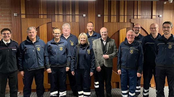 A group of eleven individuals stands together in a room, dressed in uniforms. They appear to be members of a team, possibly emergency responders or municipal workers, with a mix of genders represented. A wooden backdrop and gray carpeting provide context for their gathering.