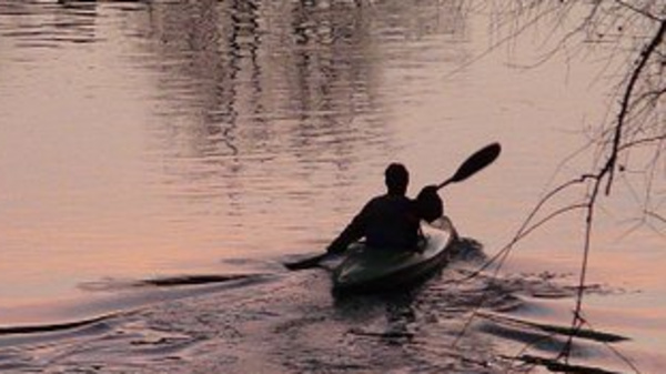 A silhouette of a person kayaking on a calm lake during sunset, with soft pink and orange reflections on the water. The surrounding scene is tranquil, highlighting the peacefulness of the outdoors.