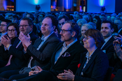Eine Gruppe von Menschen sitzt in einem gut beleuchteten Raum. Einige applaudieren, während andere aufmerksam zuschauen. Die Atmosphäre ist feierlich und fokussiert, passend zu einer Veranstaltung oder Zeremonie.