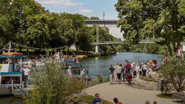 Bild eines Flusses mit Menschen, die am Ufer entspannen und Aktivitäten genießen. Im Hintergrund ist eine Brücke sichtbar. Bäume und Bootsanleger sind zu sehen. Der Text "Marbach am Neckar" und "Entdecke unsere Region" ist eingeblendet. (Text KI generiert)