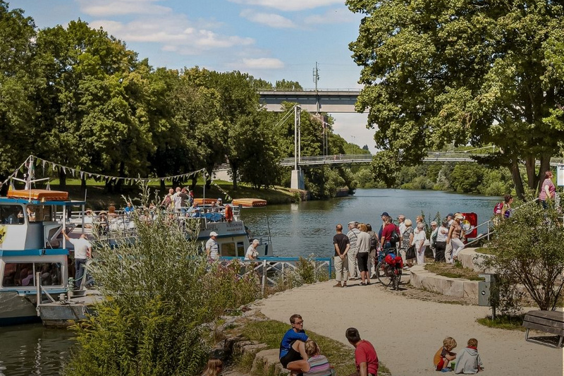 Bild eines Flusses mit Menschen, die am Ufer entspannen und Aktivitäten genießen. Im Hintergrund ist eine Brücke sichtbar. Bäume und Bootsanleger sind zu sehen. Der Text "Marbach am Neckar" und "Entdecke unsere Region" ist eingeblendet. (Text KI generiert)