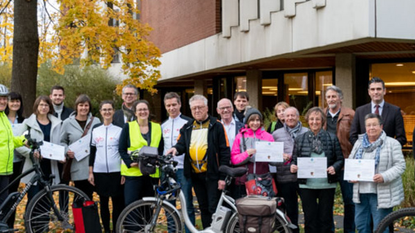 A group of individuals, including cyclists and pedestrians, stands together outside a building. They are holding certificates and wearing a mix of casual and reflective safety clothing. The setting features autumn foliage and a pathway, emphasizing a community celebration related to cycling or safety awareness.