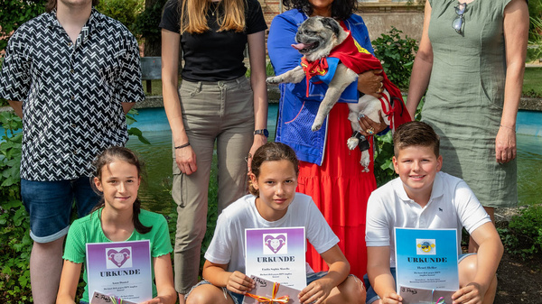 Eine Gruppe von sieben Personen steht um einen Brunnen im Freien. Die Erwachsenen stehen hinter den Kindern, die Urkunden mit dem Schriftzug "Danke" in der Hand halten. Eine Person hält einen kleinen Hund, während die anderen lächeln und in einer freundlichen Atmosphäre posieren.