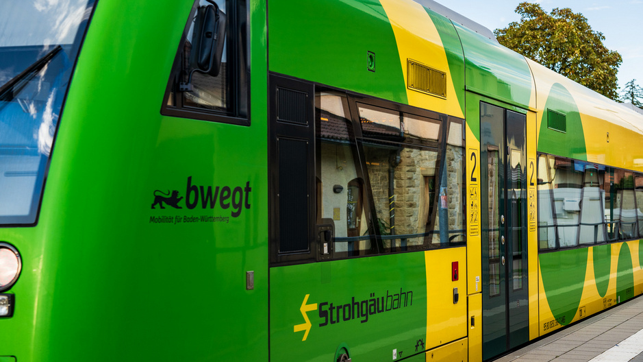 A green train with yellow accents, labeled "bwegt" and "Strohgäubahn," is parked at a station. The design emphasizes mobility and regional transportation. The platform shows a clear view of the train's side and entryway.