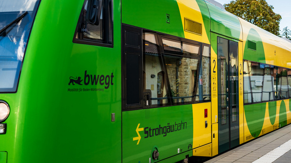 Ein grüner Zug mit gelben Akzenten steht an einem Bahnsteig. Er trägt das Logo der Strohgäubahn und die Aufschrift "bwegt" für Mobilität. Die Zugtüren sind geschlossen, und im Hintergrund sind Bäume sichtbar.