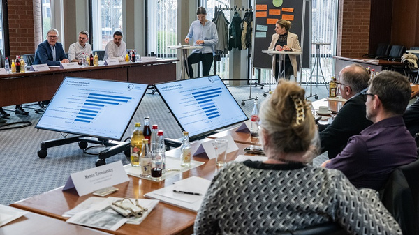 In einem Konferenzraum präsentieren mehrere Personen Daten auf großen Bildschirmen. Am Tisch sitzen Teilnehmer, die aufmerksam zuhören und Notizen machen. Im Hintergrund ist eine Pinnwand mit weiteren Informationen sichtbar. Die Atmosphäre ist professionell und diskussionsorientiert.
