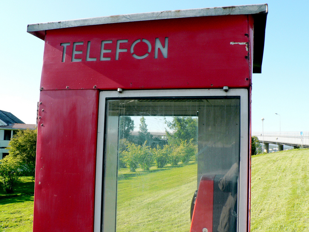 Bild: Giovanni Variottinelli, Lizenz: CC BY-NC-ND 2.0 Eine rote Telefonzelle steht auf einer Wiese. Der Schriftzug "TELEFON" ist klar und gut sichtbar an der Vorderseite angebracht. Im Hintergrund sind grüne Bäume und ein Gebäude zu erkennen.