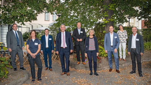A group of nine professionally dressed individuals stands outdoors, surrounded by greenery. They appear engaged and friendly, with name tags visible. The setting conveys a formal yet relaxed atmosphere, suggesting a networking or business event.